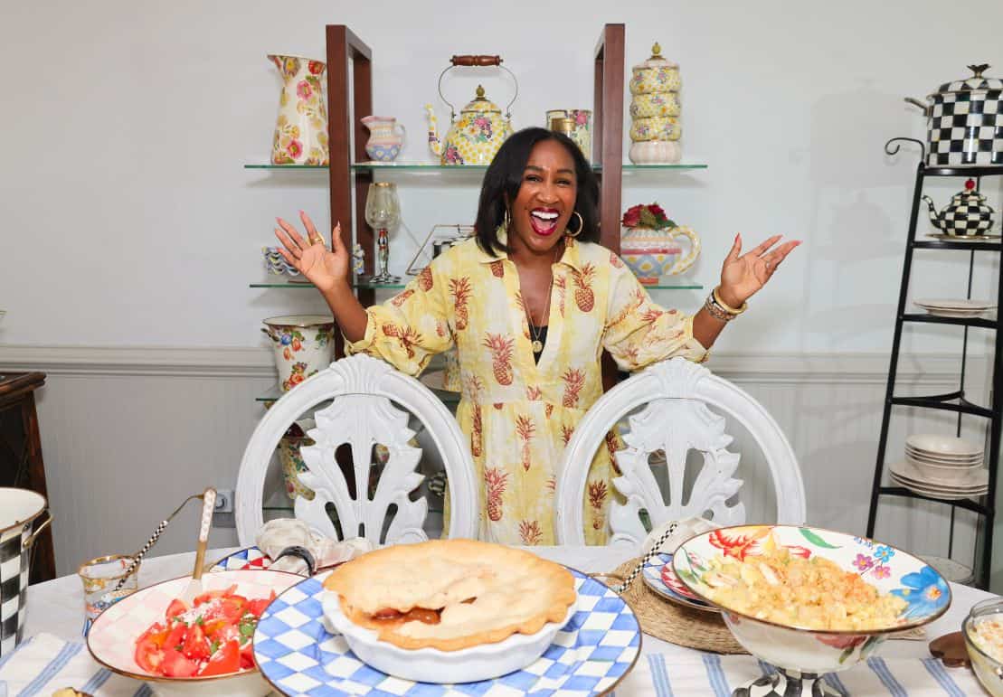 Shaunda Necole standing behind a colorful Southern holiday table with apple pie, potato salad, tomato watermelon salad, and tableware from MacKenzie-Childs for The Soul Food Pot®.
