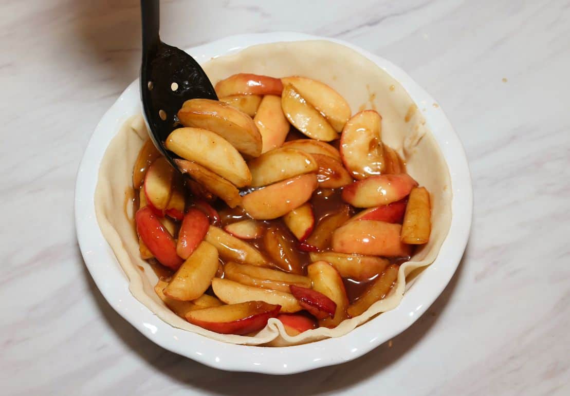 Cooked apple pie filling with thickened cinnamon juices being spooned into a bottom pie crust for The Soul Food Pot®.