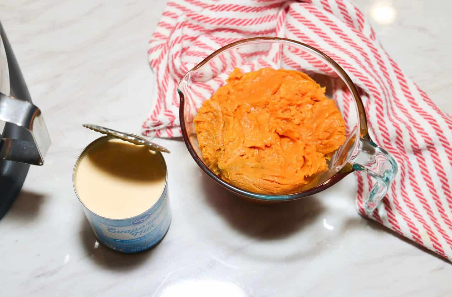 Cooked orange sweet potato flesh in a glass measuring cup beside an open can of evaporated milk, two key ingredients in The Soul Food Pot® Black Folks Sweet Potato Pie Recipe™.