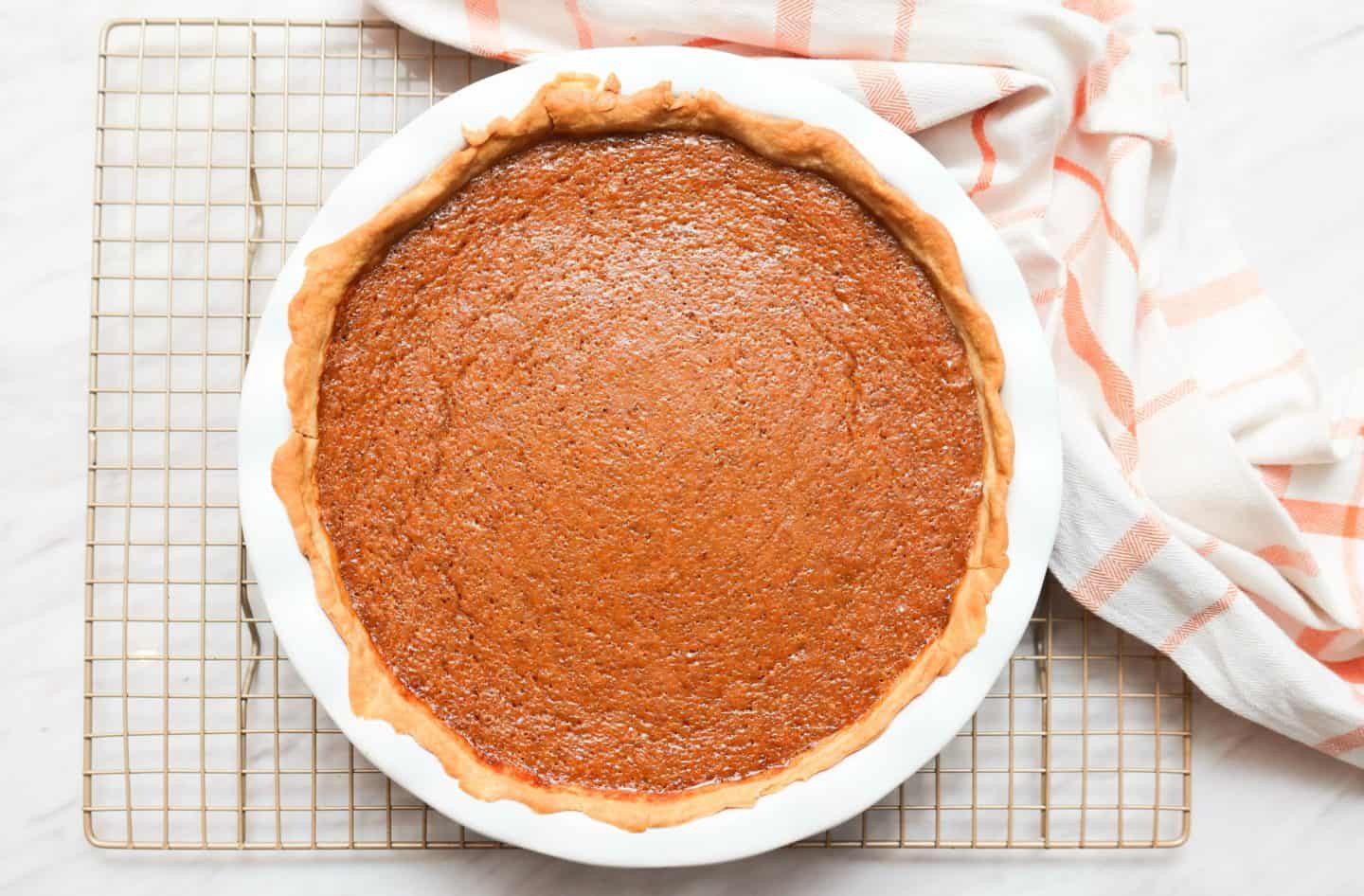 Freshly baked Black Folks Sweet Potato Pie™ in a ruffled ceramic pie dish resting on a wire cooling rack, showing its golden crust and evenly baked brown custard filling.