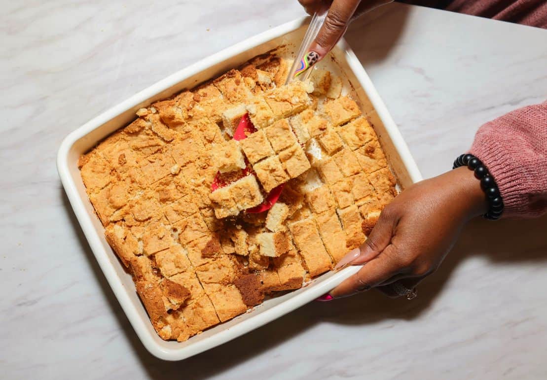 Freshly cut homemade cornbread cubes being lifted from a baking dish for The Soul Food Pot® chicken and dressing recipe.