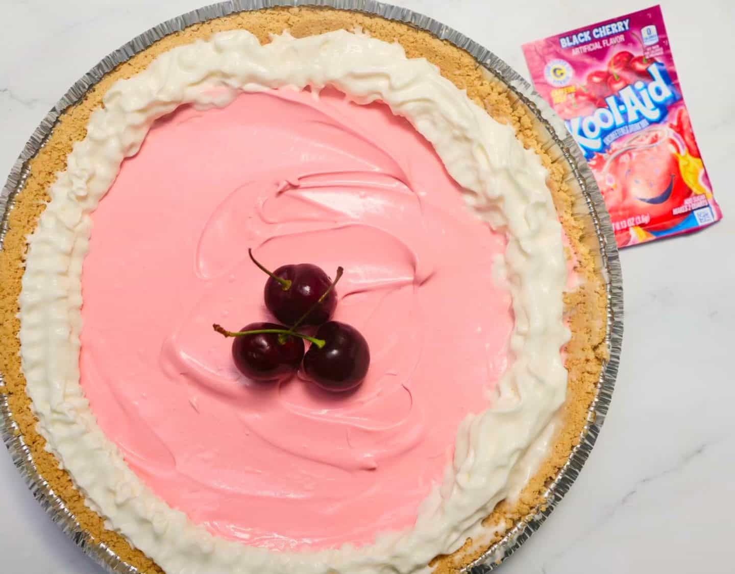 The Soul Food Pot® Black cherry Red Kool-Aid Pie with fluffy pink filling, whipped topping, fresh cherries, and a graham cracker crust, shown beside a Black Cherry Kool-Aid packet.