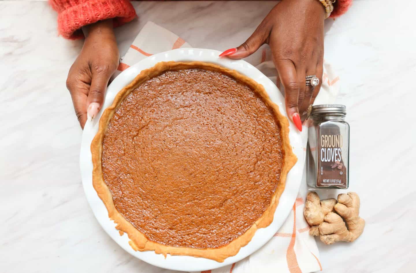 Overhead view of Shaunda Necole's Black Folks Sweet Potato Pie™ in a ruffled pie dish, showing its golden crust and baked custard filling beside fresh ginger root and ground cloves.