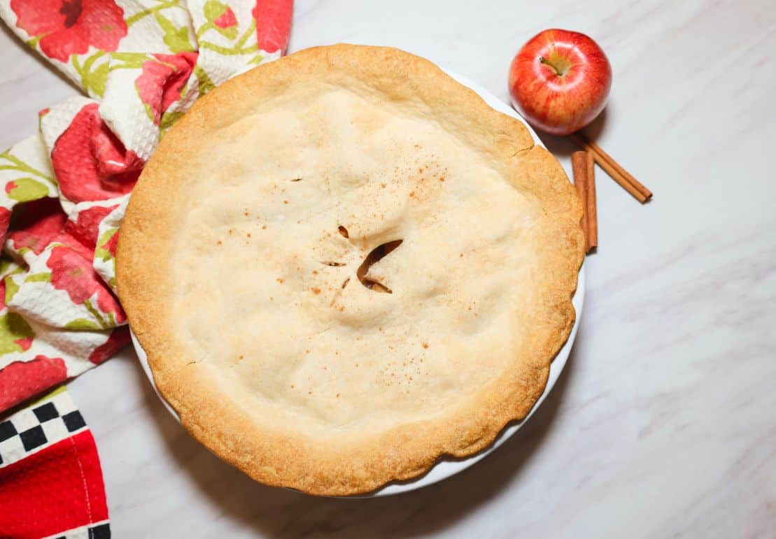 Shaunda’s Southern soul food apple pie baked in a white pie dish with a golden double crust, steam vents, red apple, and cinnamon sticks.