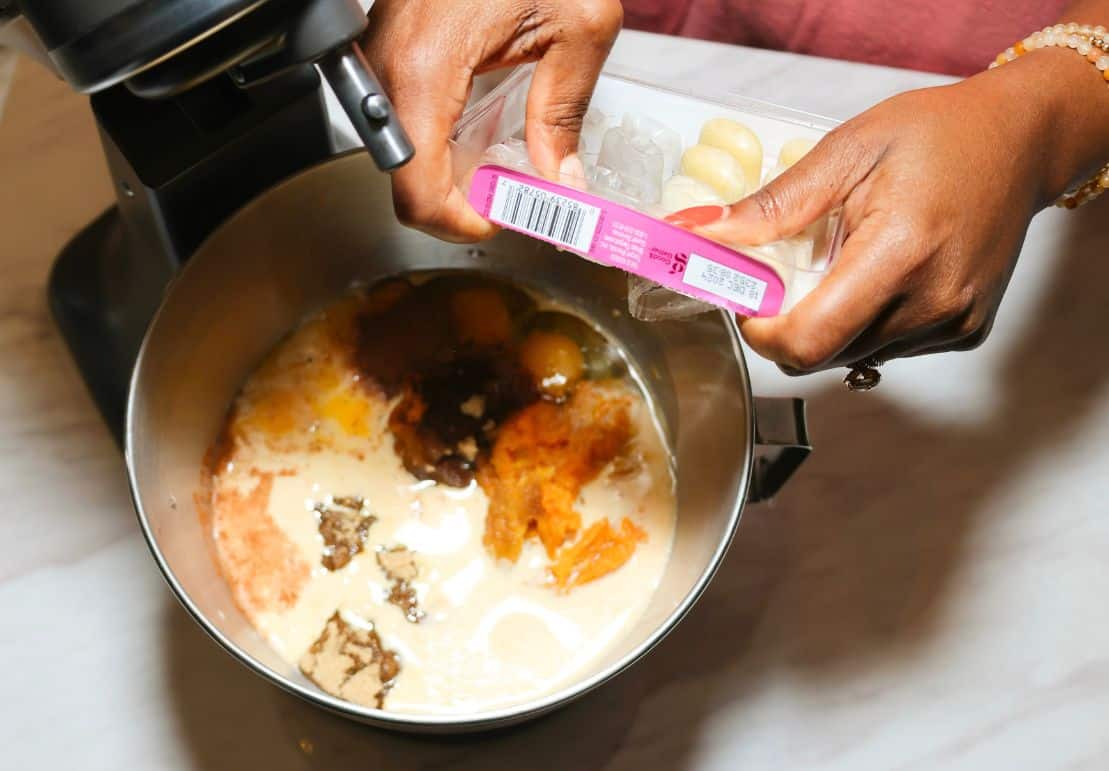 Shaunda Necole adding a frozen minced ginger cube to Black folks' sweet potato pie filling in a stand mixer bowl with mashed sweet potatoes, evaporated milk, eggs, brown sugar, and spices.