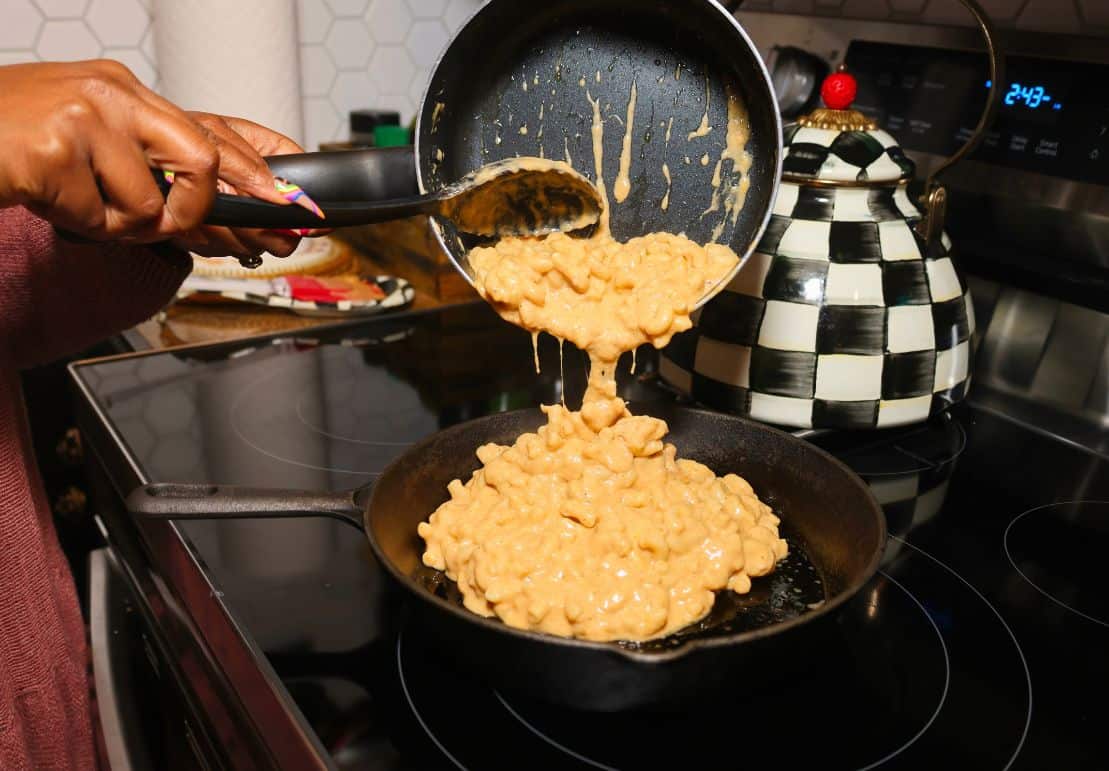 Creamy macaroni and cheese pouring from a saucepan into an 8-inch cast-iron skillet, showing the thick, cheese-coated pasta before baking.