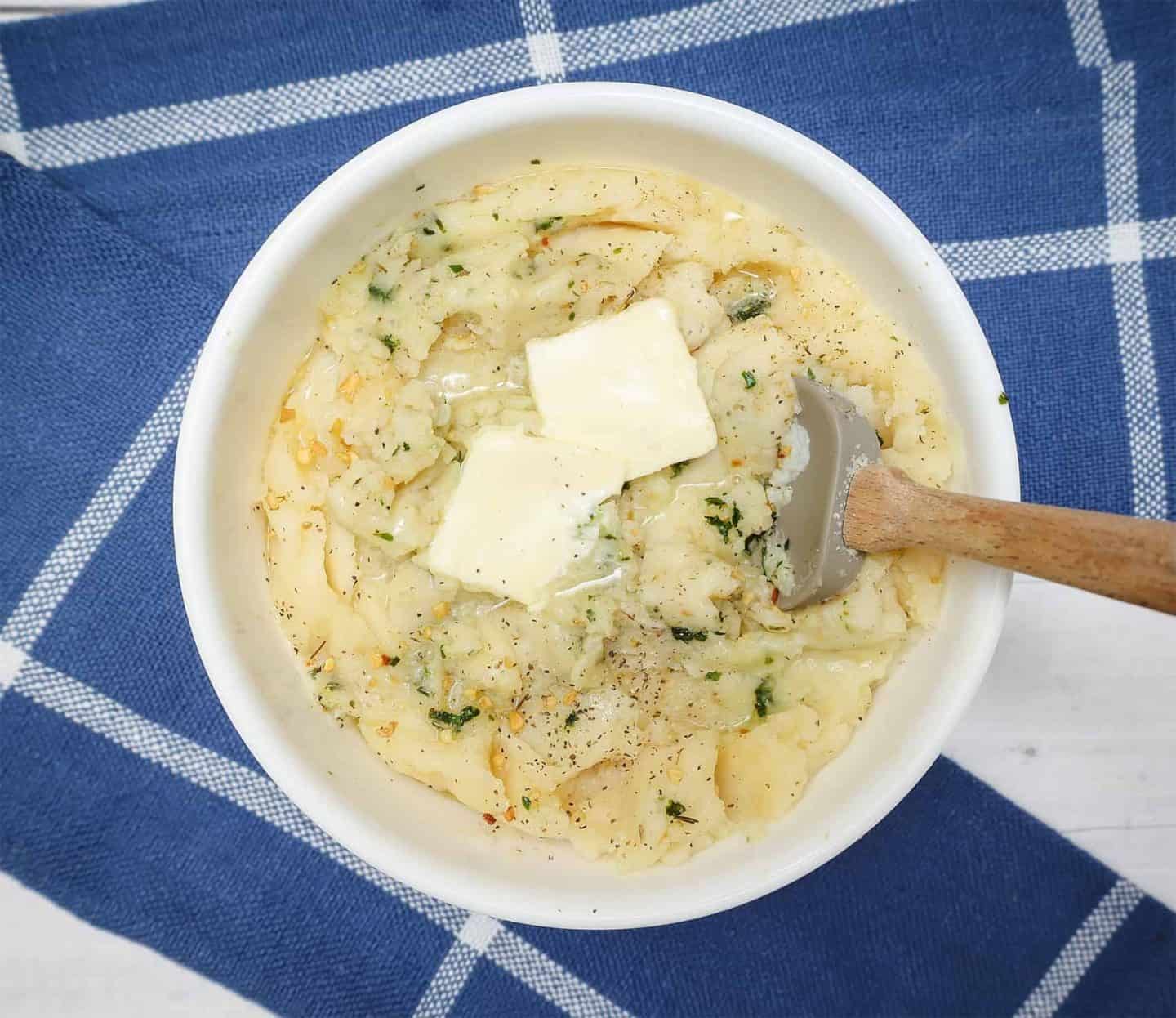 Creamy seasoned mashed potatoes topped with melting butter, herbs, and black pepper in a white bowl with a small spatula from The Soul Food Pot®.