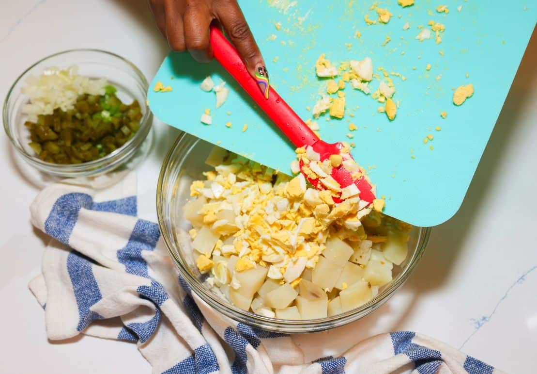 Shaunda Necole adding chopped hard-boiled eggs from a cutting board to a bowl of potatoes and dressing for her Black folks Southern potato salad recipe.