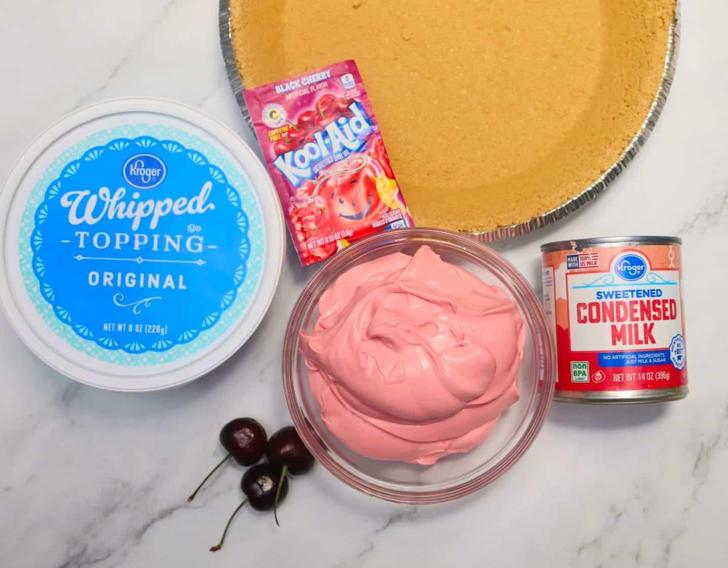 Ingredients for The Soul Food Pot® Red Kool-Aid Pie: Black Cherry Kool-Aid drink mix, sweetened condensed milk, whipped topping, prepared graham cracker crust, and fresh cherries beside the creamy pink pie filling.
