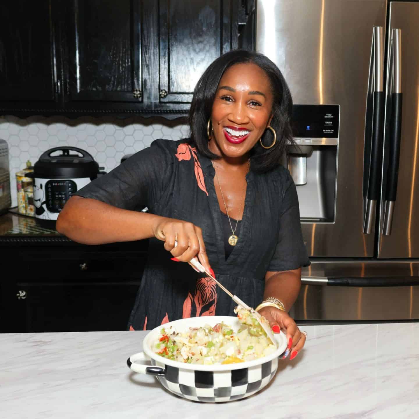 Shaunda Necole smiling while gently mixing ingredients for her Black folks Southern potato salad recipe in a black-and-white checkered bowl in her kitchen.