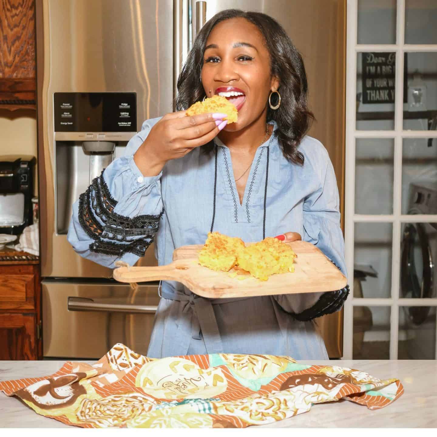 Shaunda Necole of The Soul Food Pot® tastes a piece of homemade cornbread while holding additional pieces on a wooden serving board in her kitchen.
