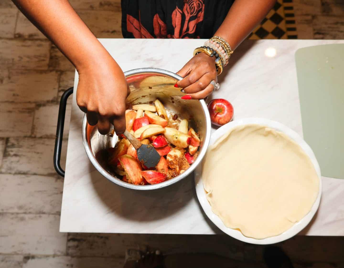 Shaunda Necole, stirring thick, unpeeled red apple slices with brown sugar and spices on the stove beside the prepared bottom crust for her Southern soul food apple pie.