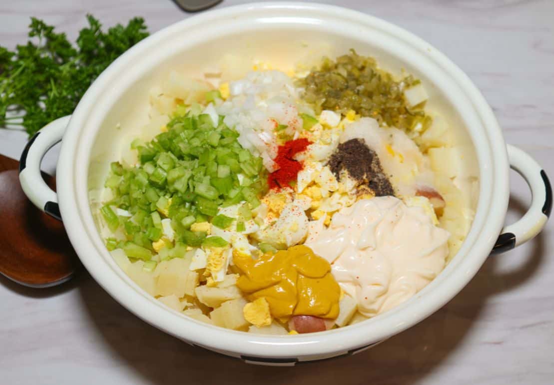 Unmixed ingredients for Shaunda Necole's Black folks Southern potato salad, with chopped potatoes, hard-boiled eggs, celery, pickles, onion, mayonnaise, mustard, and seasonings in a mixing bowl.